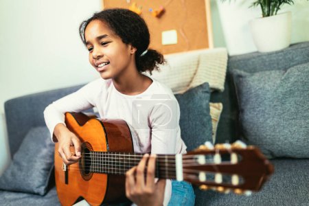 Téléchargez les photos : African american teenage girl sitting on couch in her room and learning to play guitar - en image libre de droit