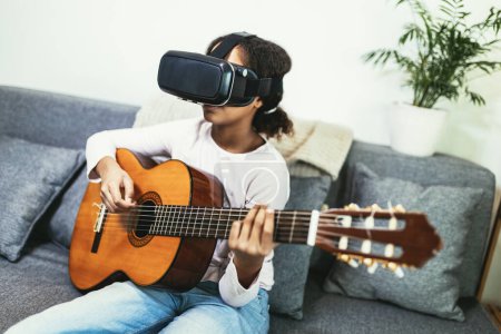Photo for African american teenage girl sitting on couch in her room and learning to play guitar using VR glasses to immerse herself in metaverse - Royalty Free Image