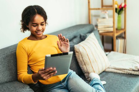 Téléchargez les photos : Happy African young girl using digital tablet technology sitting on couch at home. Smiling young girl using apps, make video call, browsing internet on sofa. - en image libre de droit