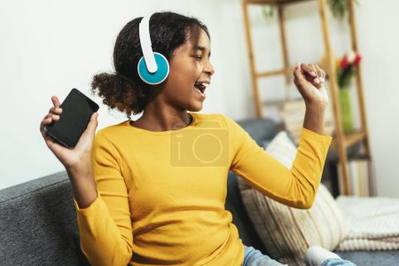Téléchargez les photos : Afro-américaine profiter de temps libre à la maison tout en étant assis sur le lit et en écoutant de la musique avec des écouteurs - en image libre de droit