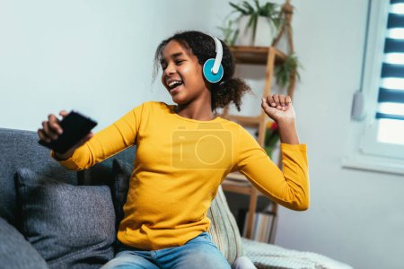 Téléchargez les photos : African American girl enjoying free time at home while sitting on the bed and listening to music with headphones - en image libre de droit