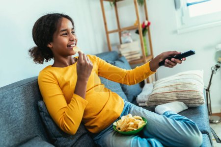 Téléchargez les photos : Enfant regardant la télévision assis sur le canapé. Jeune fille afro-américaine à la maison et elle regarde la télévision - en image libre de droit