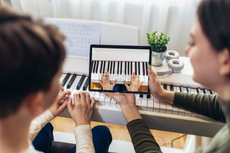 Photo for Brother and sister play electric piano at home and have fun. The sister helps her younger brother to play piano.They use a digital tablet for online lessons. - Royalty Free Image