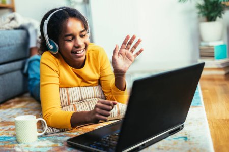 Foto de Escuela sonriente chica afroamericana en los auriculares viendo la lección de vídeo en la computadora, niño feliz en los auriculares tienen clase web en línea utilizando el ordenador portátil en casa, concepto de educación en casa - Imagen libre de derechos