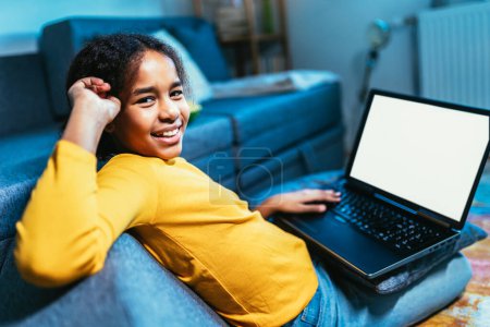 Téléchargez les photos : École souriante fille afro-américaine regarder la leçon vidéo sur ordinateur, enfant heureux ont classe web en ligne à l'aide d'un ordinateur portable à la maison, concept d'enseignement à domicile - en image libre de droit