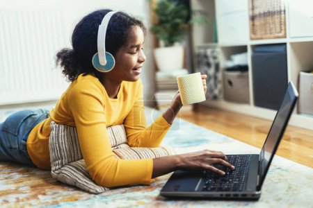 Photo for Smiling school African American girl in headphones watching the video lesson on computer,  happy child in earphones have online web class using laptop at home, homeschooling concept - Royalty Free Image