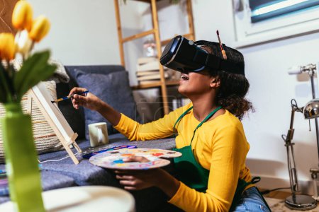 Téléchargez les photos : Happy black school girl painting with a paintbrush at home using VR glasses to immerse herself in the metaverse - en image libre de droit