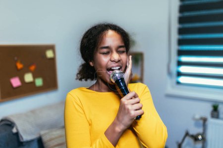 Téléchargez les photos : Jolie préadolescente noire tenant un microphon chantant du karaoké à la maison, enregistrant des chansons pour concours. Concept de style de vie des enfants - en image libre de droit