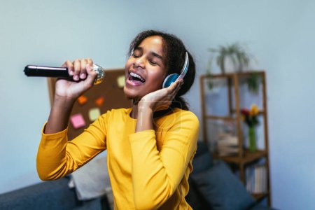 Photo for Cute preteen black girl  holding microphon singing karaoke at home, recording songs for contest. Children's lifestyle concept - Royalty Free Image