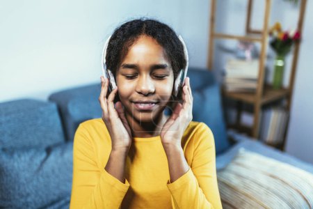 Téléchargez les photos : Afro-américaine profiter de temps libre à la maison tout en étant assis sur le lit et en écoutant de la musique avec des écouteurs - en image libre de droit