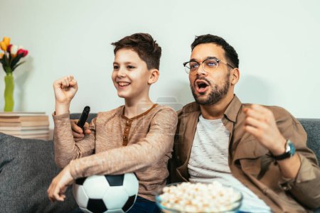 Téléchargez les photos : Garçon regardant match de football avec son père à la maison, assis sur le canapé - en image libre de droit