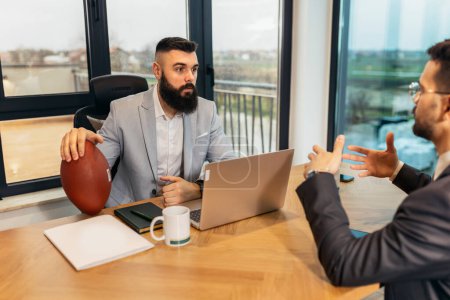 Photo for Handsome young businessman holding a rugby ball in the office when talking with his colleague - Royalty Free Image