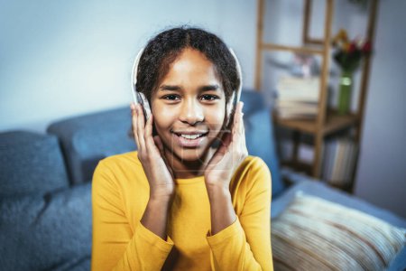 Foto de African American girl enjoying free time at home while sitting on the bed and listening to music with headphones - Imagen libre de derechos