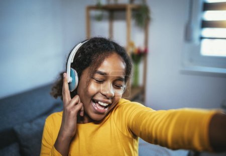 Photo for African American girl enjoying free time at home while sitting on the bed and listening to music with headphones - Royalty Free Image
