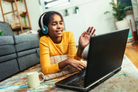 Téléchargez les photos : Ecole souriante afro-américaine fille dans les écouteurs regarder la leçon vidéo sur ordinateur, enfant heureux dans les écouteurs ont classe web en ligne à l'aide d'un ordinateur portable à la maison, concept d'enseignement à domicile - en image libre de droit