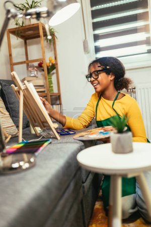 Foto de Black preteen girl happily at a table, surrounded by an array of creative materials for painting to inspire their next great idea. - Imagen libre de derechos