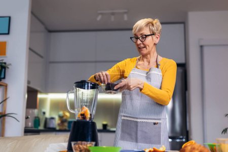 Photo for Senior woman in casual home clothes prepares healthy cocktails with different seasonal fruits. - Royalty Free Image