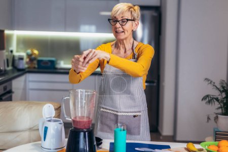Foto de Mujer mayor en ropa casera casual prepara cócteles saludables con diferentes frutas de temporada. - Imagen libre de derechos