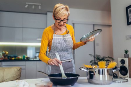 Téléchargez les photos : Heureuse femme âgée cuisine dans sa cuisine moderne - en image libre de droit