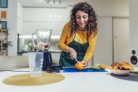 Photo for Beautiful, happy young woman in casual home clothes prepares fitness cocktails with different seasonal fruits. - Royalty Free Image