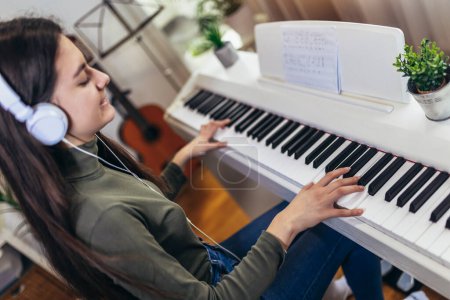 Téléchargez les photos : Fille heureuse joue du piano pour son passe-temps temps de détente dans le salon à la maison. Portrait d'adolescente souriante à la maison jouant du piano - en image libre de droit