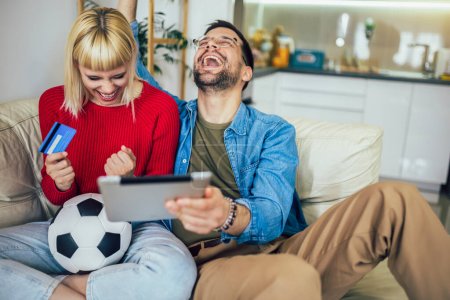Pareja joven viendo un partido de fútbol en una tableta digital y usando una tarjeta de crédito para apostar en línea.