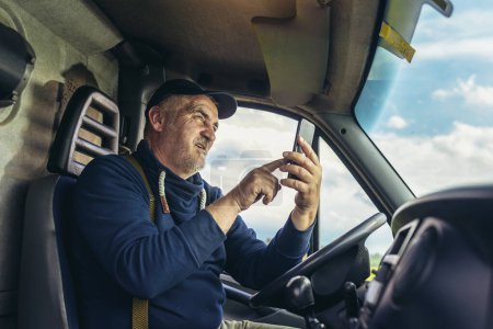 Conductor de camión maduro utilizando el teléfono móvil mientras conduce el vehículo de transporte.