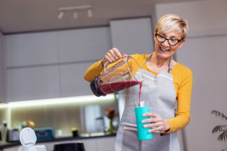Photo for Senior woman in casual home clothes prepares healthy cocktails with different seasonal fruits, drinking juice and having fun. - Royalty Free Image
