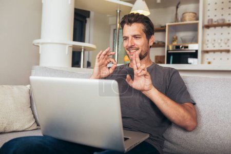 Téléchargez les photos : Jeune homme montrant un geste en langue des signes à l'aide d'un ordinateur portable, faire un appel vidéo. - en image libre de droit