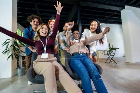 Foto de Jóvenes empresarios alegres en ropa casual inteligente divirtiéndose mientras compiten en sillas de oficina y sonriendo - Imagen libre de derechos