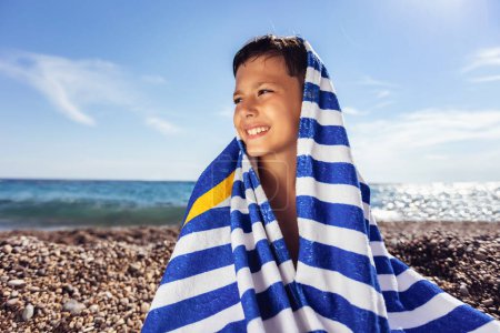 Foto de El niño en la playa cubierto con una toalla de playa a rayas azul y blanco y sonriendo felizmente. - Imagen libre de derechos