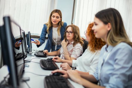 Téléchargez les photos : Un groupe d'étudiants s'assoient devant un bureau dans un laboratoire informatique. Ils sont tous concentrés sur leurs écrans que leur professeur féminin fait son chemin autour de vérifier sur chaque élève individuellement. - en image libre de droit