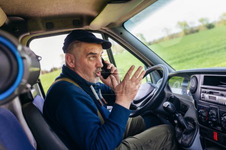 Photo for Mature truck driver using mobile phone while driving transport vehicle. - Royalty Free Image