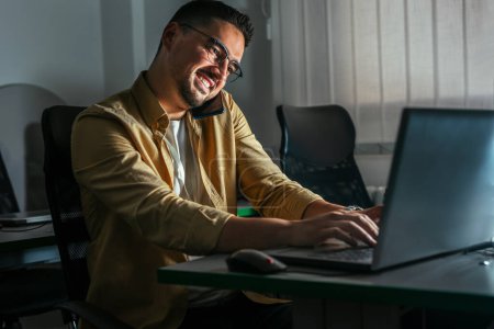 Photo for Young man working late in the office. A man is sitting at a laptop in a dark office and using a smart phone. - Royalty Free Image