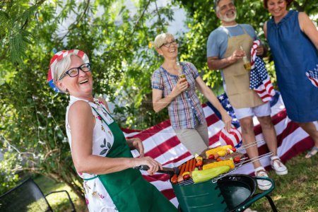 Photo for A group of elderly people celebrates the 4th of July in a backyard. They are making barbeque, vegetables, drinking beverages, making jokes, and laughing. - Royalty Free Image
