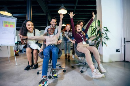 Photo for Multiethnic coworkers racing in their chairs while on a break in their office. They are laughing and enjoying their jo - Royalty Free Image
