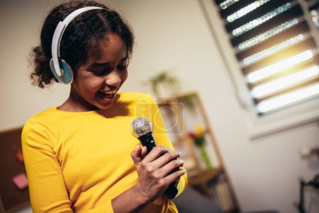 Téléchargez les photos : Jolie préadolescente noire tenant un micro chantant du karaoké à la maison, enregistrant des chansons pour un concours. Concept de style de vie des enfants - en image libre de droit