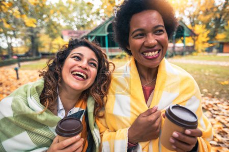 Foto de Dos jóvenes amigas sonrientes charlando al aire libre y tomando café mientras disfrutan del paseo por el parque juntas - Imagen libre de derechos