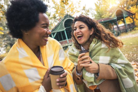 Téléchargez les photos : Deux jeunes femmes souriantes amis bavardant à l'extérieur et buvant du café tout en profitant de la promenade dans le parc ensemble - en image libre de droit
