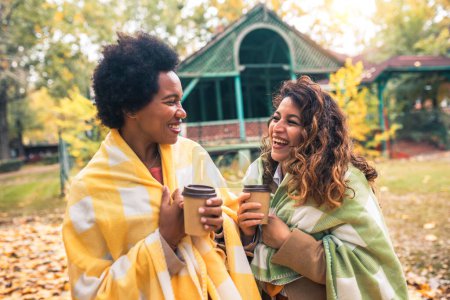Téléchargez les photos : Deux jeunes femmes souriantes amis bavardant à l'extérieur et buvant du café tout en profitant de la promenade dans le parc ensemble - en image libre de droit