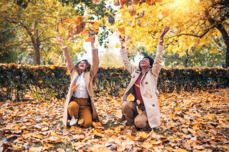 Photo for Two smiling young women friends throwing leaves in the park full of fall colors. They are laughing and having fun - Royalty Free Image