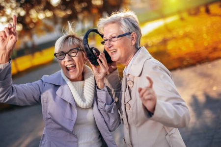 Photo for Two older women listening to music on their wireless headphones in the colorful park in the autumn - Royalty Free Image