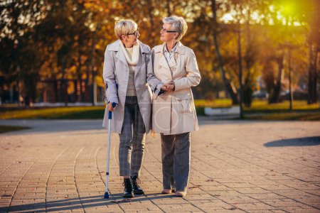 Téléchargez les photos : Une femme plus âgée aide une autre femme qui marche avec une canne dans le parc coloré en automne tout en souriant et en faisant des blagues - en image libre de droit