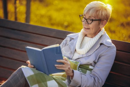 Foto de Una mujer mayor leyendo un libro. Ella está sentada en el banco en el colorido parque en el otoño - Imagen libre de derechos