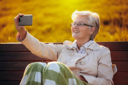 Téléchargez les photos : Une femme plus âgée prend des selfies assis sur le banc dans le parc coloré à l'automne - en image libre de droit