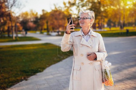 Photo for An older woman walking in the park and taking pictures of it with her groceries in the autumn - Royalty Free Image