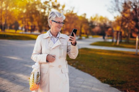 Téléchargez les photos : Une femme plus âgée se promène dans le parc coloré et regarde son téléphone transporter ses courses à l'automne - en image libre de droit