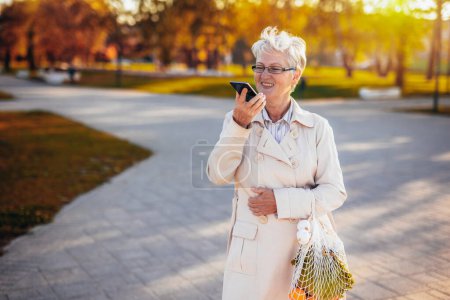 Foto de Una mujer mayor caminando por el parque y enviando un mensaje de voz mientras llevaba sus comestibles en el otoño - Imagen libre de derechos