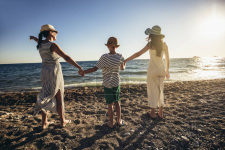 Téléchargez les photos : Mère et enfants se tenant la main profitant du coucher de soleil sur la plage. Happy concept de voyage et de vacances en famille. - en image libre de droit