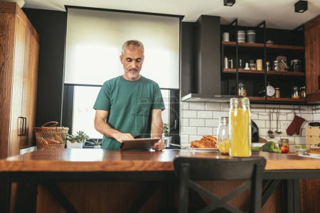 Téléchargez les photos : Homme mûr dans la cuisine travaillant à partir de la maison en utilisant une tablette numérique - en image libre de droit